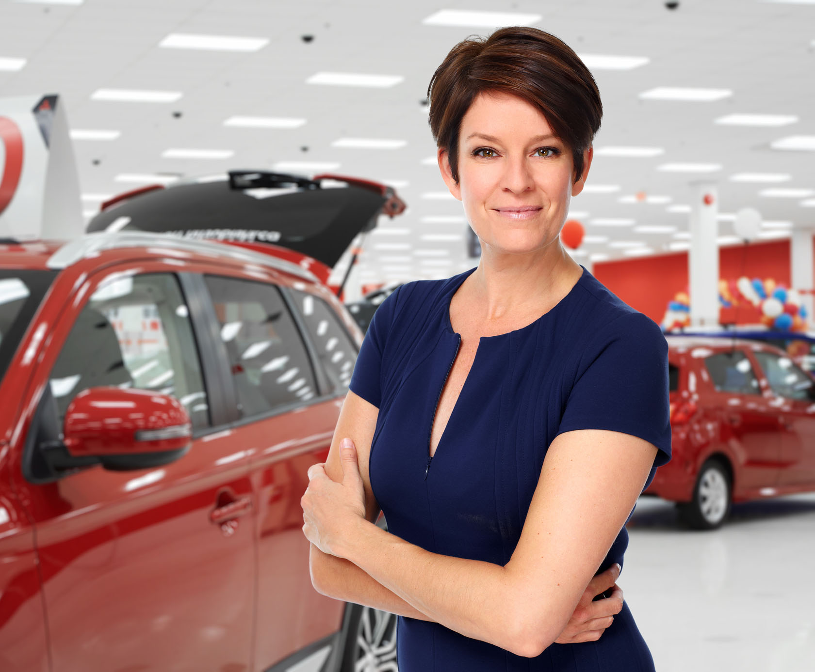 A confident woman in a navy dress stands with arms crossed in a vibrant car dealership, showcasing various red vehicles in the background.